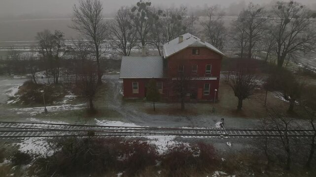 Aerial views old red brick railway station beside single track line in winter morning fog. Vntage rural transit scene with atmospheric mood for travel and documentary use.
