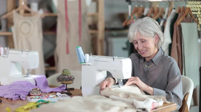 Attentive mature female dressmaker working with sewing machine in tailor's workshop