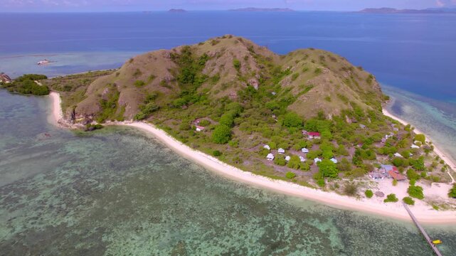 Backward aerial footage of Kanawa Island in Indonesia showcasing private yachts and boats anchored near a wooden jetty, with small beachfront cottages and huts along a pristine white sand beach.