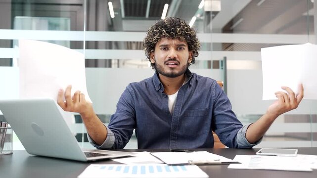 Confused disappointed businessman having difficulty with paperwork sitting at desk at workplace in business office. Puzzled worker checks documents and cannot understand problem and looking in camera