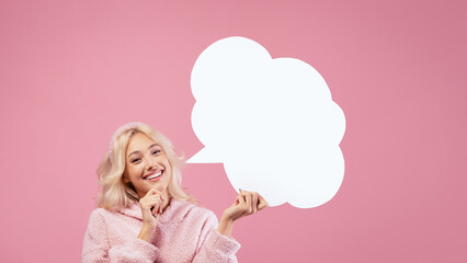 A woman stands against a pink background. She is smiling and holding a blank speech bubble with one hand. She has light hair and wears a cozy outfit. © Prostock-studio