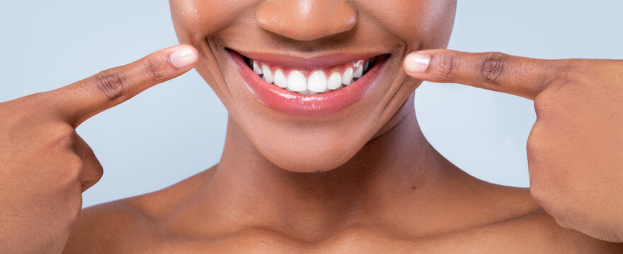 A woman is showing her bright smile while pointing at her teeth with her fingers. She is indoors against a light background. Her joy and positivity are clear in her expression.