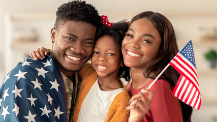 A smiling family stands close to each other while holding an American flag. The father wraps himself in a star-spangled blanket, and the daughter and mother smile brightly. © Prostock-studio