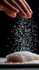Fototapeta premium Hand sprinkles coarse salt over a raw fish fillet, creating a cascade of salt crystals in mid-air. The scene suggests a culinary preparation in progress with focus on texture