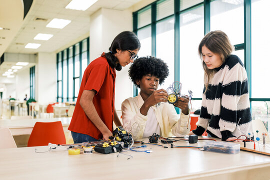 Three students collaborate to assemble a small wheeled robot in a bright classroom, surrounded by electronic components and tools.