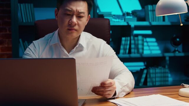 Overload Asian tired Korean business man with laptop reading documents incorrect data at evening office middle-aged Chinese businessman working online bankruptcy problem insomnia headache at night