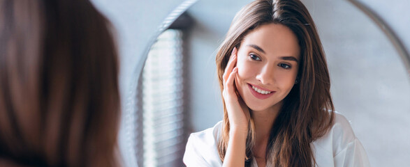 A woman stands in front of a mirror with a smile on her face. She is touching her cheek as she checks her appearance. The setting is bright and simple, capturing a moment of self-care. © Prostock-studio