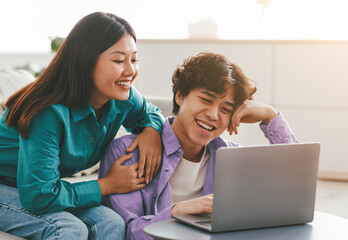 Fototapeta premium People and technologies concept. Happy loving young Asian couple using laptop, watching video or movie, shopping remotely, checking social media online, relaxing in living room