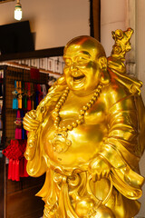 Fototapeta premium A golden laughing Buddha statue stands prominently inside a shop. The statue wears a large necklace and holds a cane. Colorful items hang in the background, showing a lively market scene