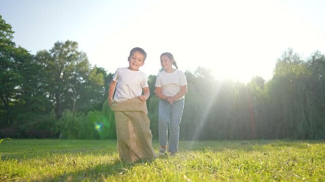 Boy jumps in sack race on sunny field while girl runs behind. Children play outdoor game with burlap sack. Kids compete in race on grass. Boy hops in sack as girl cheers. Children enjoy sunny game.