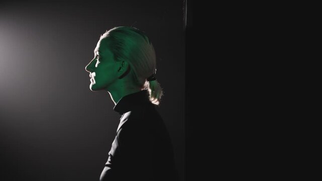 A young blonde woman with a short ponytail makes a step to the left, entering a beam of light. High-contrast side lighting sculpts her pensive profile, capturing the essence of overcoming silence and 