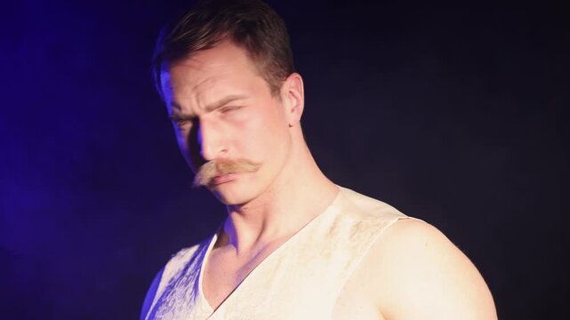 A charismatic muscular man with a mustache, captured in profile, flinches as a powerful spotlight hits his face, quickly raising his hand to block the light. Close-up.