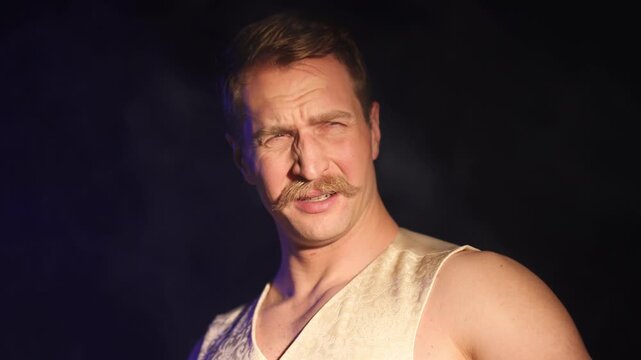 A charismatic, muscular man with a mustache stands thoughtfully in profile until a bright light suddenly illuminates his face, causing him to react in fear and shield himself with his hand. Close-up.