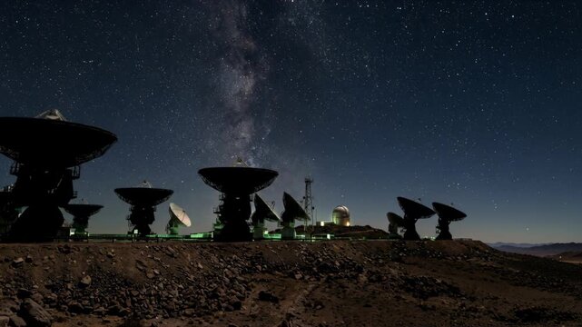 Radio telescope array under starry night sky with Milky Way, rocky foreground and remote observatory antenna emitting faint green lights