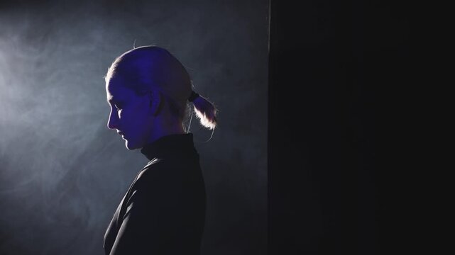 A young blonde woman with a ponytail takes a left step into a dramatic side light. High-contrast video emphasizes her contemplative profile, representing the moment of finding one's voice and facing t
