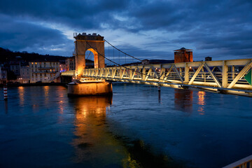 Fototapeta premium Illuminated pedestrian bridge over the Rhône river at night in Vienne, France. Beautiful night view of the Saint-Romain-en-Gal footbridge with golden lights reflecting in the water.