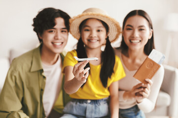 Fototapeta premium Smiling young girl in hat hold plane, selective focus. Millennial japanese parents happy and show passport and tickets in living room interior, blurred. Family vacation, travel and holiday together