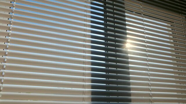 Bright morning sunlight shining through white horizontal window blinds as sun moves across blue sky, creating shifting shadows, light patterns on slats in cozy home interior