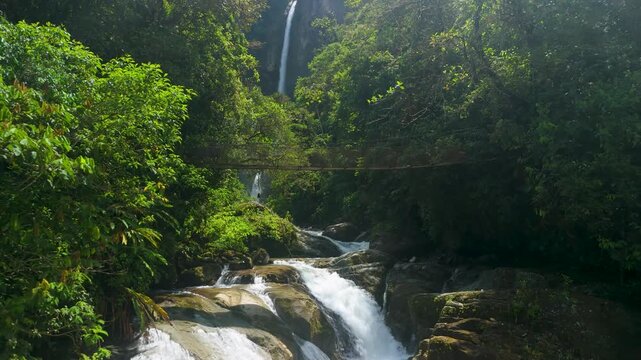 Camera tilts up from rushing whitewater rapids and rocky riverbed to reveal wooden suspension bridge with Cascada El Rocio Machay waterfall plummeting through lush green tropical rainforest canopy