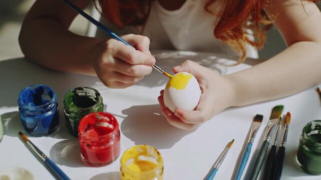 Young girl with red hair paints a white egg with yellow color using a brush, surrounded by jars of paint and brushes on a bright table, Easter concept