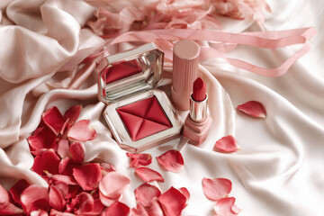 Red lipstick and a compact with a matching shade sit on smooth fabric adorned with rose petals. 