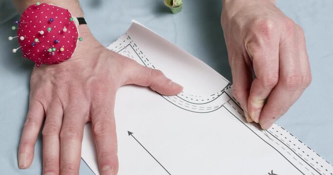 Professional tailor uses thin metal sewing pins to securely fasten paper pattern onto fabric laid out on table. Man attaches pattern to soft textile surface