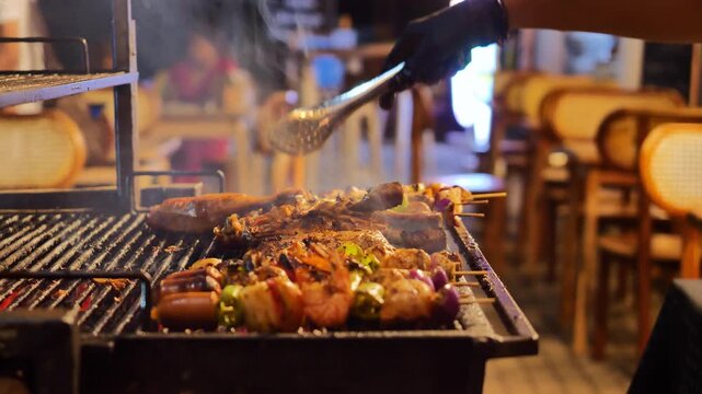 Barbecue grill with shrimp sausage kebab and steak cooking over charcoal, chef hand turning skewers with tongs, smoky outdoor dinner and summer party concept
