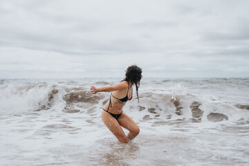 Fototapeta premium A woman in a black bikini splashes through shallow surf on a cloudy beach, conveying carefree summer energy, outdoor fitness, and coastal relaxation.