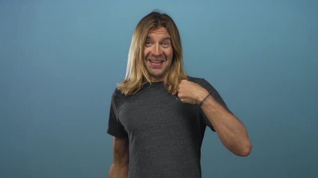 Man with long blond hair wearing a casual dark t shirt points finger to chest and smiles in blue studio; identity confidence.