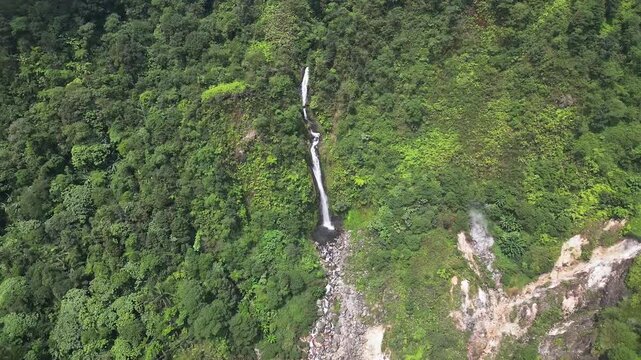 Drone aerial landscape featuring Curug Cikawah waterfall flowing into rocky terrain amid dense green rainforest in Indonesia. Scenic tropical environment inside Gunung Halimun Salak National Park.