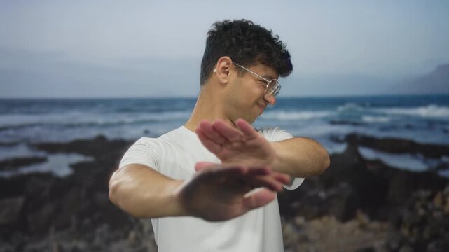 Young hispanic man wearing glasses shields face with both arms on rocky shoreline beach; avoidance discomfort.