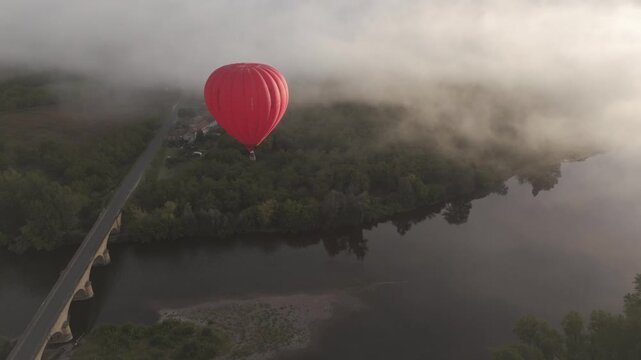 Aerial view of a red hot air balloon drifts serenely above a bridge and river, with mist adding an ethereal quality to the landscape, Castelnaud-la-Chapelle, France.