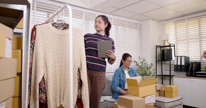 Asian women small business partners checking clothing inventory, coordinating online selling tasks, preparing online shopping parcel box together inside home based e-commerce warehouse