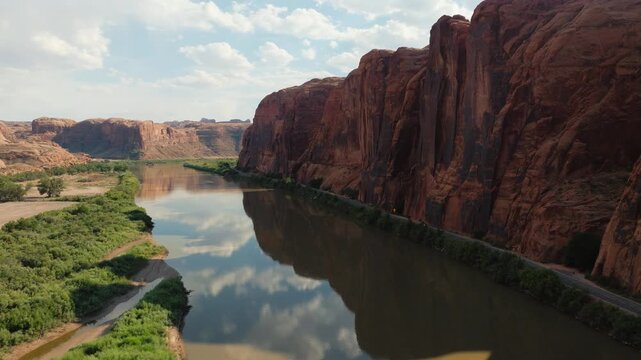 Aerial view of the Colorado River reflecting the sky, flowing between towering red rock cliffs and green vegetation, Moab, Utah, United States.