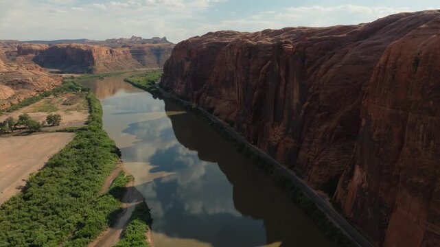Aerial view of the winding Colorado River reflecting the sky as it cuts through towering red cliffs and lush green vegetation, Moab, Utah, United States.
