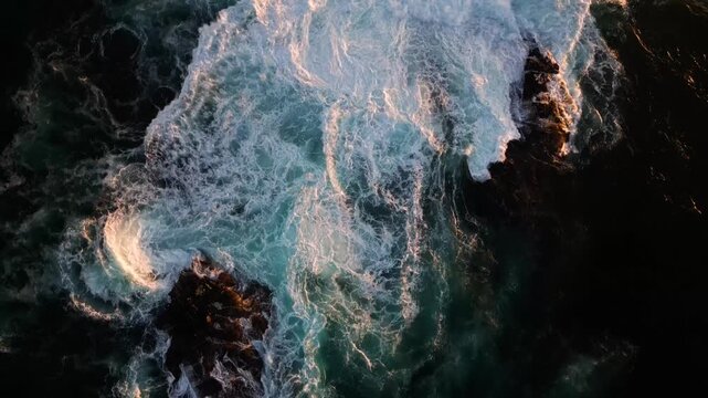 Aerial view of intense ocean waves crashing against dark, rugged rocks, creating a mesmerizing display of contrasting colors and textures, Cape Town, Western Cape, South Africa.