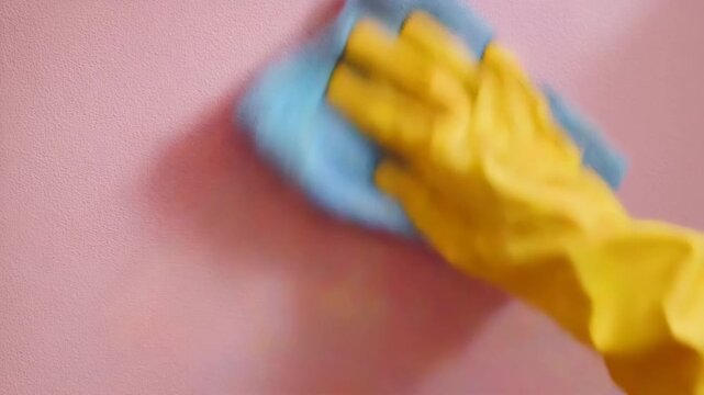 close up of hand in yellow rubber glove wiping pink wall with blue microfiber cloth