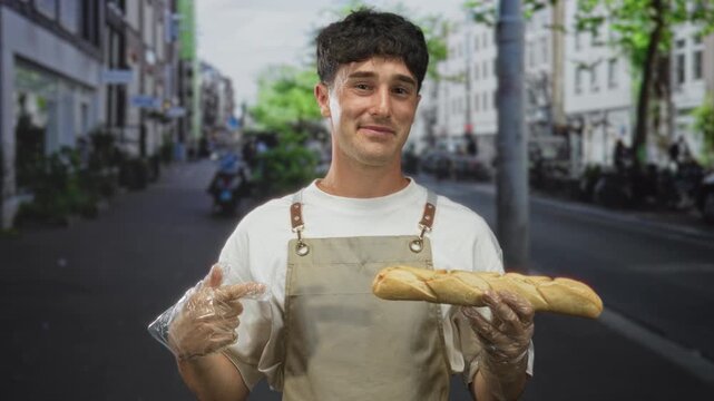 Man holding a baguette and points finger at bread on street while wearing apron and plastic gloves among city storefronts; friendly service.