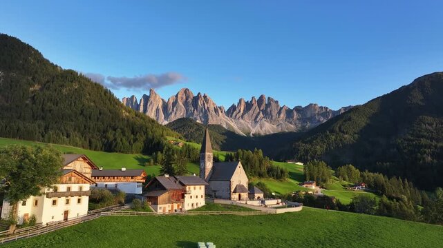 Aerial view of Santa Magdalena Church nestled among green hills, with the majestic Dolomites mountains as a backdrop, Santa Magdalena, Trentino-Alto Adige, Italy.