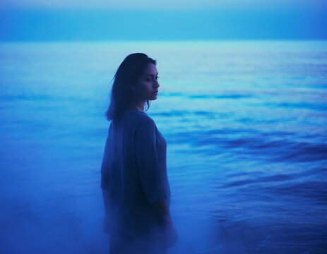 A single person standing near calm ocean water in the early morning, surrounded by soft blue mist.
