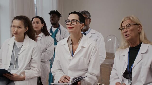 Multiethnic team of healthcare professionals dressed in white coats listening to unknown speaker and taking notes while attending research conference