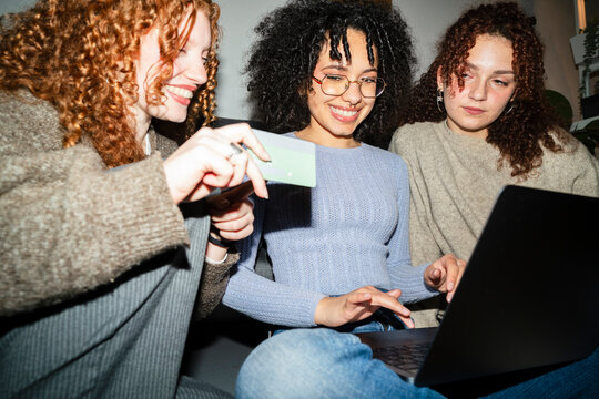 Three young women with curly hair are gathered around a laptop, one holding a card, smiling and engaged in online activity.