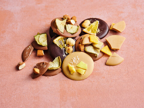 A beautiful pile of assorted chocolate discs featuring dark, milk, and caramel varieties, topped with crunchy hazelnuts, almonds, dried apple, orange, and mango pieces on a pink surface