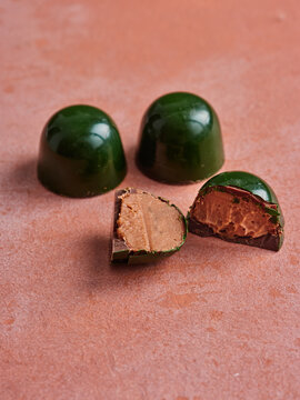 Three elegant dark green chocolate bonbons rest on a textured pink surface. Two bonbons remain whole, while one is cut open, revealing its rich, creamy brown filling inside
