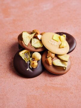 A cluster of decadent chocolate discs featuring dark, milk, and white chocolate varieties. Each disc is garnished with an array of dried fruits and crunchy nuts, presented on a vibrant pink surface