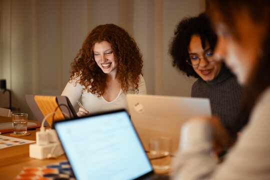 Three young women collaborate on a project at a table, using laptops and a tablet. One woman with curly red hair smiles brightly as she works.