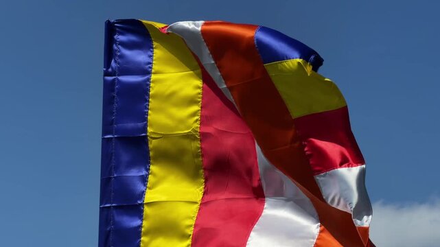 Buddhist flag with vertical colored stripes moves steadily in the breeze against a bright open sky in a cultural outdoor setting. Spiritual tradition.