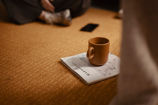 A person sits on a textured floor with a mug resting on a magazine. A mobile phone is visible in the background.
