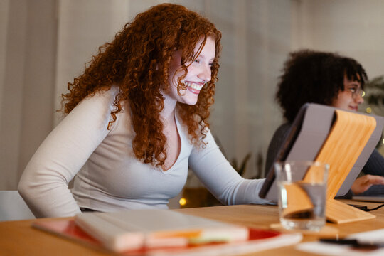 A young woman with curly red hair smiles while looking at a tablet. Another person is visible in the background, also working on a device.