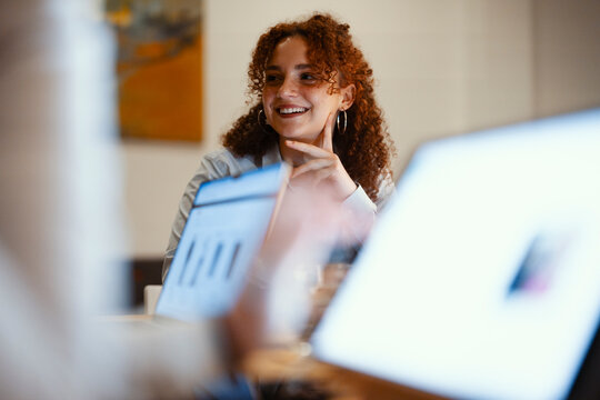 A young woman with curly hair smiles while looking at a laptop screen, engaged in a collaborative work session with colleagues.